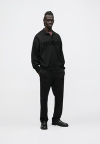 Juodas sportinis kostiumas, sudarytas iš megztinio su "HUGO" logotipu ir atitinkančių kelnių, pagamintų iš minkštos medžiagos. Aptakus dizainas, derinamas su elegantiškais juodais batais.