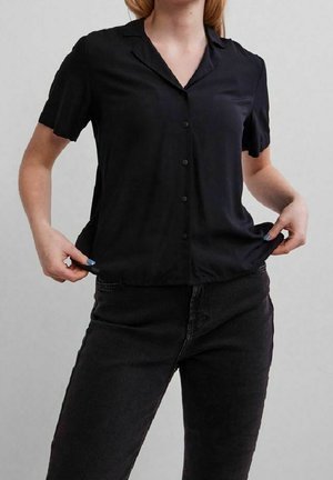 Kobieta w czarnej koszuli na krótki rękaw z guzikami i czarnych dżinsach, trzymająca obie ręce na dolnym brzegu koszuli.