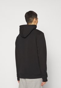 Sudadera negra con capucha ajustable, textura suave, mangas largas y puños acanalados. Lleva puesta sobre pantalones grises claros, mostrando un ajuste casual.