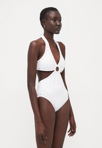 Costume da bagno intero bianco con scollo all'americana, taglio frontale con un accento circolare e dettagli con borchie lungo i lati.