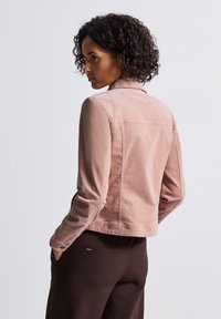 Kort rosa jacka med en framträdande krage, tillverkad av mjukt tyg. Har en figurnära design och sidofickor, kombinerad med mörka byxor.