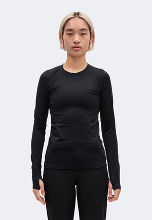 Le mannequin se tient face à l’objectif sur un fond uni, vêtu d’un haut noir ajusté à manches longues et d’un pantalon noir.