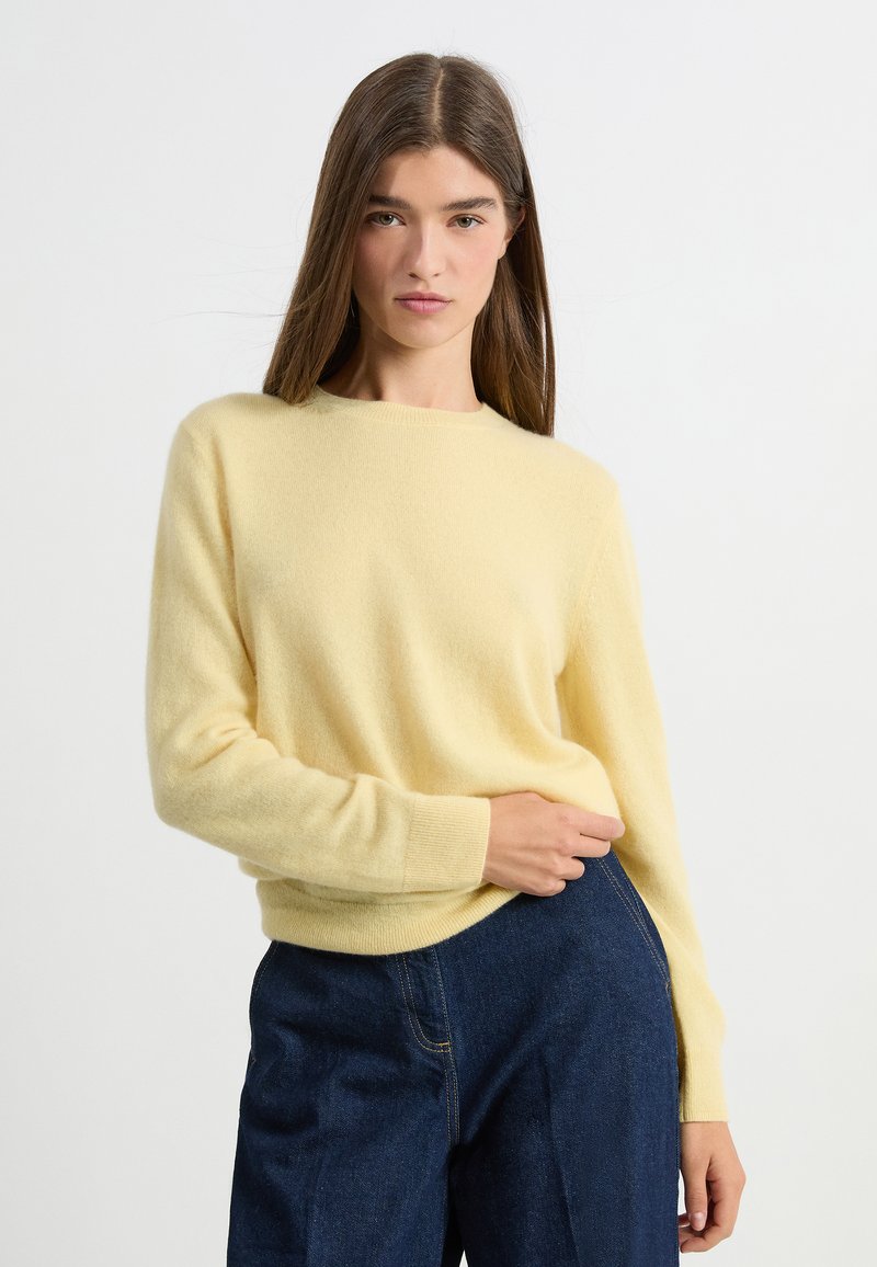 Żółty, miękko wyglądający sweter dzianinowy z długimi rękawami i ściągaczami, noszony z ciemnoniebieskimi spodniami na jednolitym tle.