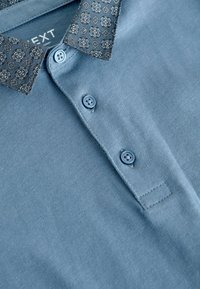 Blaues Poloshirt mit einem gemusterten schwarzen Kragen, zwei Knöpfen, weichem Griff und einem kleinen Stoffetikett in der Nähe des Halsausschnitts.