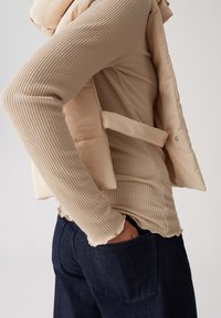 Beige gerippter Langarmshirt, das unter einer hellbeigen Steppweste getragen wird, kombiniert mit dunkelblauen Jeans, die über eine Gesäßtasche verfügen.
