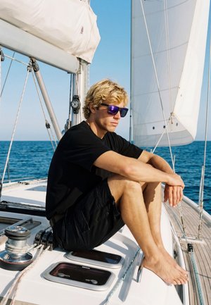 Jeune homme en chemise noire et short portant des lunettes de soleil, assis pieds nus sur le pont d'un voilier avec la mer et les voiles visibles en arrière-plan.