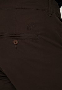 Mörkbruna byxor med en framficka som har knäppning och präglad logga. Tyget har en slät textur och klassisk design.