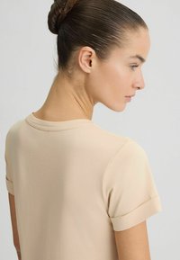 Beige t-shirt met korte mouwen, een ronde hals en omgeslagen mouwen. Zachte stof met een gladde textuur, wat de eenvoudige uitstraling benadrukt.