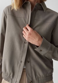 Grijze button-up jas met een gestructureerde afwerking, voorzien van een kraag, zichtbare knopen en elastische manchetten. Draagt over een lichtgekleurde ribbeltop.