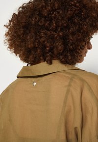 Personne aux cheveux bouclés vue de dos, portant une chemise transparente marron clair avec un petit bouton métallique rond sur le haut du dos.