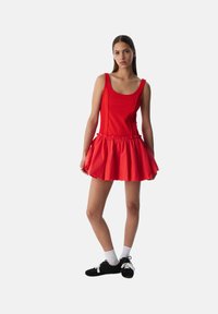 Rode jurk met een aansluitend lijfje en een uitlopende rok, met een geplooide zoom. Gemaakt van een gladde stof, gecombineerd met zwarte schoenen en witte sokken.