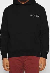 Svart huvtröja med känguruficka, med en vit "HILFIGER"-logotyp på vänster bröst. Materialet verkar mjukt, designen är klassisk med dragsko i huvan.