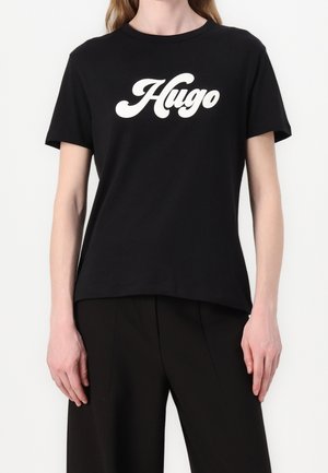 Person trägt ein schwarzes kurzärmeliges Shirt mit weißem kursivem "Hugo"-Text und schwarze Hosen, steht vor einem einfarbigen Hintergrund.