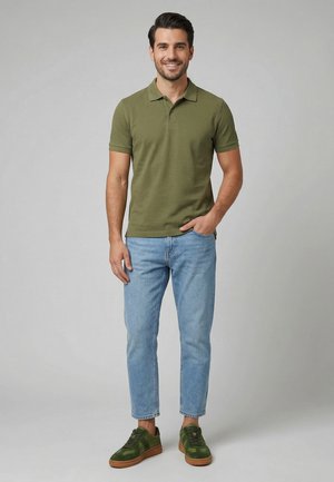 Lächelnder Mann steht mit Hand in der Hosentasche, trägt ein olivgrünes Polo, hellblaue Jeans und olivgrüne Sneakers vor einem einfarbig grauen Hintergrund.