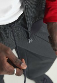 Svarta sportiga joggers med ett texturerat tyg, elastisk midja och dragsko. En kontrasterande röd jacka med dragkedja syns ovanpå.