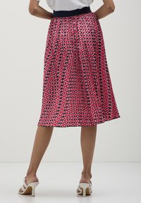 Donna che indossa una gonna midi plissettata con motivi geometrici rosa, rossi e neri e sandali alti bianchi, in piedi contro uno sfondo neutro.