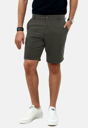 Mann in olivgrünen Shorts, schwarzem Shirt, weißen Sneakers und Armbanduhr, der mit einer Hand in der Hosentasche vor einem weißen Hintergrund steht.