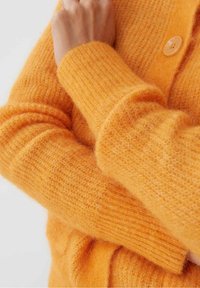 Orange stickad tröja med en mjuk, texturerad yta. Har ribbade ärmmuddar och en rund knappdetalj.