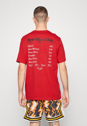 Mężczyzna w czerwonej koszulce z napisem „Breakfast Club” i listą treningów na plecach oraz w krótkich spodenkach w czarno-pomarańczowo-biały wzór, stoi tyłem na jednolitym tle.