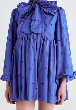 Fialové šaty s dlouhým rukávem zdobené květinovým vzorem s očky, volánkovými manžetami a velkou mašlí uvázanou u krku.