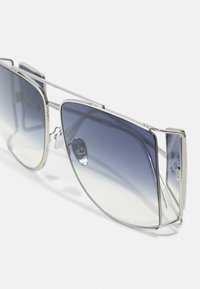 Occhiali da sole in metallo argentato con lenti oversize che presentano una gradazione dal blu scuro al trasparente. Design minimalista con linee eleganti e puntini sul naso.