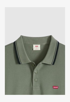 Oliwkowa zielona koszulka polo wykonana z fakturowanej tkaniny, z klasycznym kołnierzykiem, dwoma guzikami oraz granatowymi paskami na krawędziach kołnierza. Czerwona naszywka z logo.