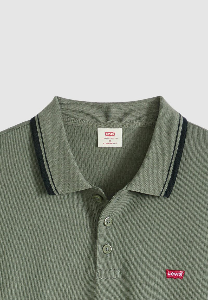 Polo color verde oliva realizzata in tessuto strutturato, con colletto classico, due bottoni e righe blu navy ai bordi del colletto. Toppa con logo rosso.