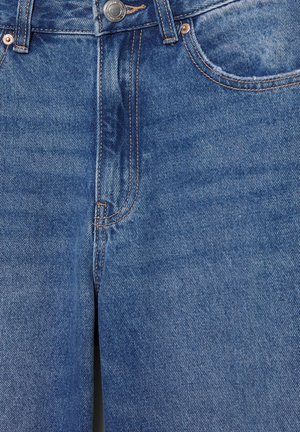 Kék farmernadrág fém gombos záródással, látható első zsebekkel és narancssárga kontrasztos varrással közeli felvételben.