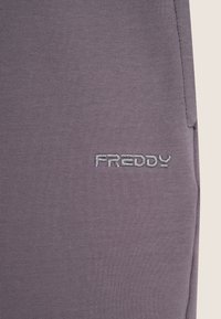 Pantalon de jogging violet en tissu doux, arborant un logo "FREDDY" brodé en argent à l'avant, avec une texture lisse.