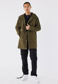 Olijfgroene waterproof jas met een capuchon en drukknoopsluitingen, gecombineerd met zwarte cargobroeken en witte sneakers, staand op een witte achtergrond.