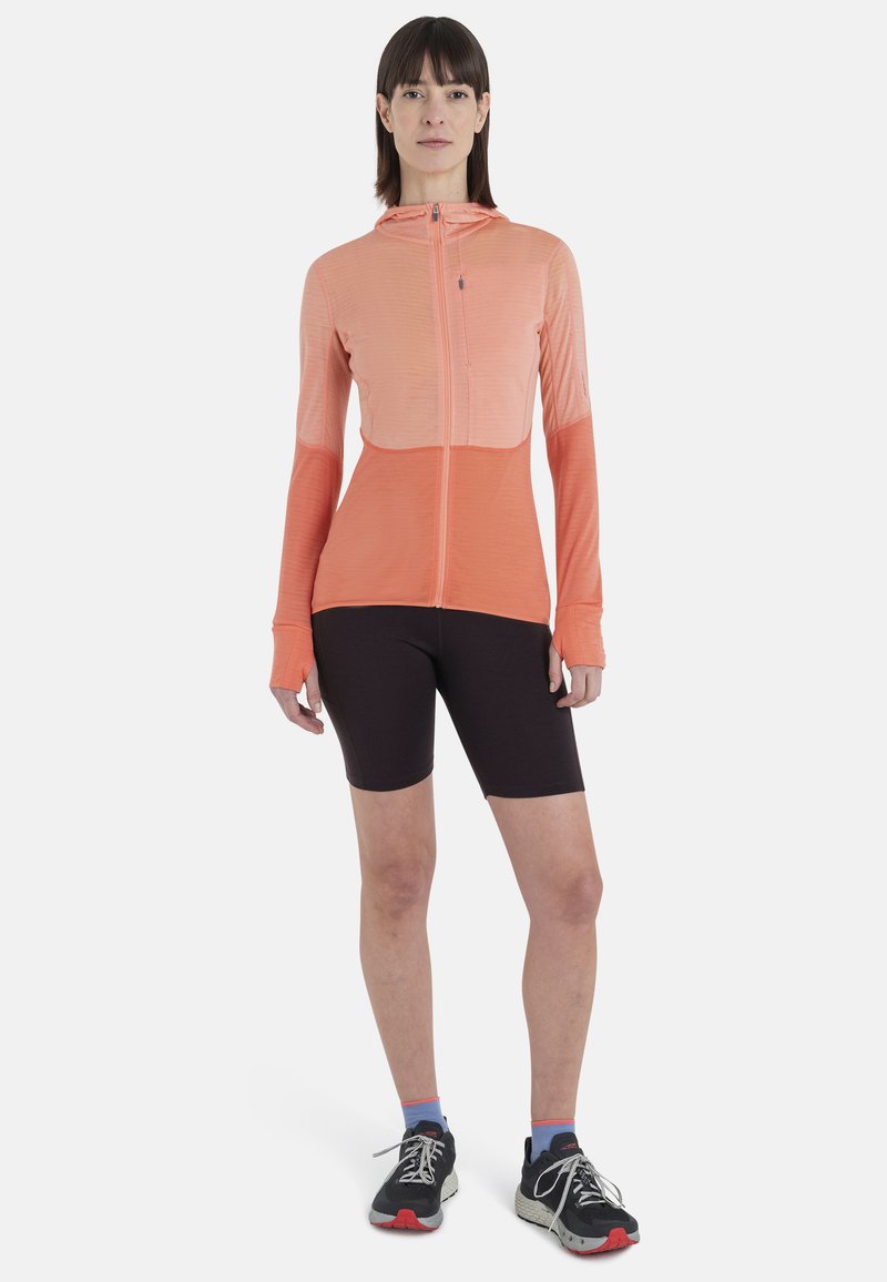 Femme debout portant une veste à capuche zippée corail, un short de cyclisme noir, des chaussettes bleues et des chaussures de course noires sur un fond blanc.