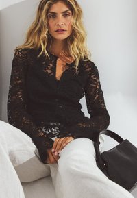 Black lace cardigan with long sleeves and button front, paired with light-colored linen trousers and a small black handbag.