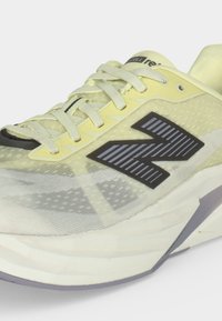 Κοντινή λήψη ανοιχτόκίτρινου παπουτσιού New Balance για τρέξιμο με μαύρο λογότυπο, λευκά κορδόνια και μαλακή λευκή σόλα σε γκρι φόντο.