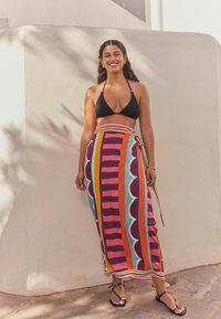 Zwarte bikini-top met een kleurrijke, gemusterd wikkelrok met gedurfde strepen en geschulpte accenten. Platte sandalen maken de look compleet.