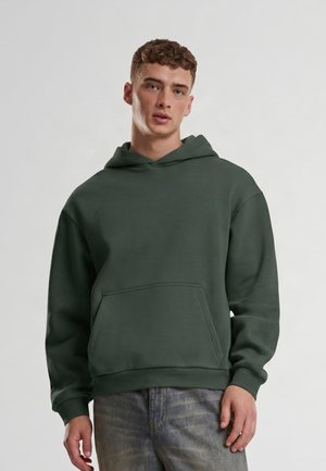 Jeune homme aux cheveux courts bouclés portant un sweat à capuche vert foncé et un jean clair délavé, debout devant un fond clair uni.