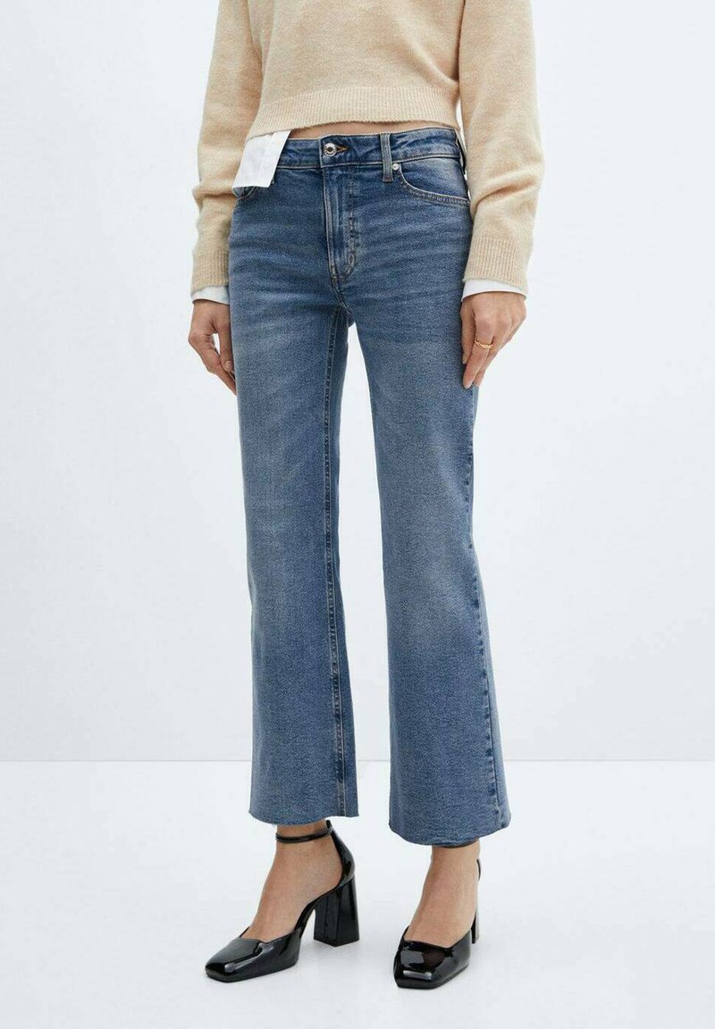 Vrouw in cropped blauwe jeans, beige trui, witte shirtmanchetten en zwarte gepatineerde leren enkelschoenen met enkelband, staande tegen een effen achtergrond.