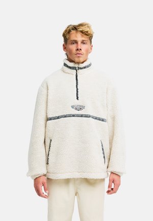 Blonder Mann trägt einen weißen Fleecepullover mit Viertelreißverschluss und gemustertem Saum, steht vor einem einfarbigen hellen Hintergrund.