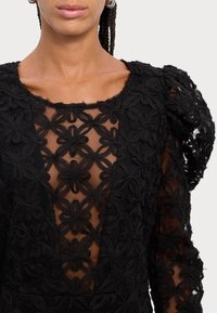 Svart blommig spetsklänning med en genomskinlig kropp med intrikata blommönster och strukturerade puffärmar. Djup V-ringning.
