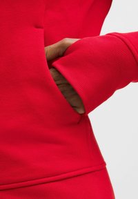 Červená mikina s klokaní kapsou, žebrovanými manžetami a hladkým povrchem. Detail záběru ukazuje ruku sahající do kapsy.