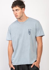 Camiseta de algodón azul claro con cuello redondo y mangas cortas. Presenta un estampado de ave y follaje en color azul marino en el lado izquierdo del pecho.