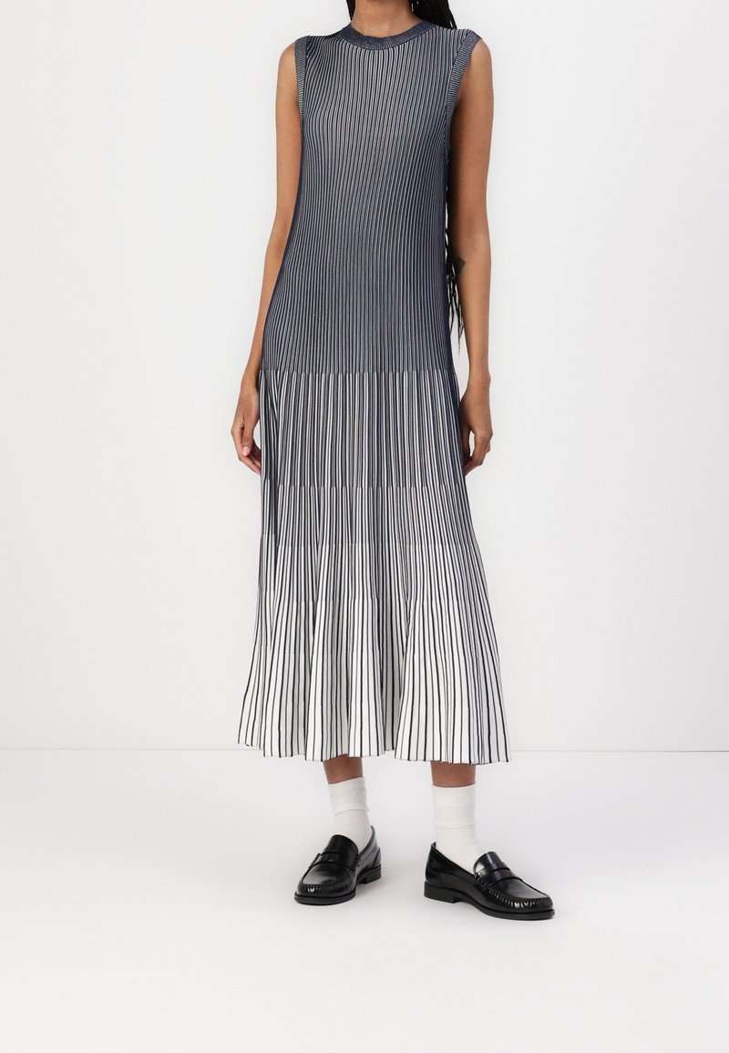 Robe midi plissée sans manches à rayures verticales noires et blanches, associée à des chaussettes blanches et des mocassins noirs.