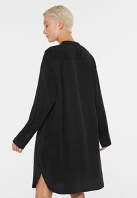 Schwarzes Hemdkleid aus weichem Stoff, mit langen Ärmeln, einem kragenlosen Ausschnitt und seitlichen Schlitzen für mehr Bewegungsfreiheit.