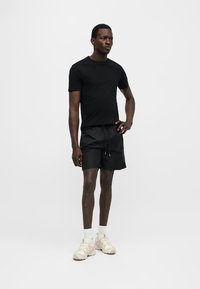 Άνδρας μοντέλο στέκεται μπροστά σε λευκό φόντο φορώντας μαύρο μπλουζάκι, μαύρα σορτς με κορδόνι, λευκές κάλτσες και μπεζ αθλητικά παπούτσια.