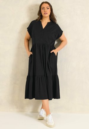 Femme debout en robe midi noire à volants avec manches courtes et poches, portant des baskets blanches, sur un fond clair uni.