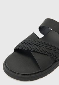 Black sandal featuring a woven strap design, flat sole, and two wide bands. Made of synthetic material with a smooth texture.
