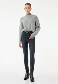 Suéter de cuello alto de canalé gris, jeans oscuros de cintura alta y botas negras hasta el tobillo con un tacón cuadrado. Diseño simple, silueta ajustada.