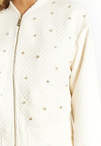 Cremefarbene Bomberjacke aus strukturiertem Stoff mit einem Rautenmuster, akzentuiert durch kleine goldene und kristallene Verzierungen sowie einen Reißverschluss.