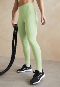 Jasno zielone, wysokie legginsy wykonane z elastycznego materiału, zestawione z czarnymi butami sportowymi z białymi akcentami i teksturowaną podeszwą.