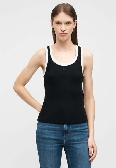 Γυναίκα που φοράει μαύρη αμάνικη ριπ μπλούζα με λευκή επένδυση και μπλε τζιν, στέκεται με το ένα χέρι στη μέση μπροστά σε ένα απλό φόντο.