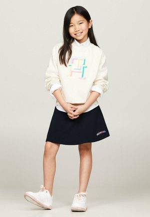 Sudadera blanca con un logo multicolor, combinada con una falda azul marino. Zapatillas blancas con acentos rosas, el modelo se presenta con las manos entrelazadas.